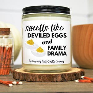 May include: A white candle jar with a black lid and a label that reads "Smells like Deviled Eggs and Family Drama" with a picture of deviled eggs. The candle is on a wooden surface with a matchbox and a pumpkin.