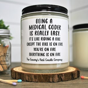 May include: A white candle with a black lid and a black label that reads "Being a Medical Coder is Really Easy. It's like riding a bike except the bike is on fire. You're on fire. Everything is on fire. The Canary's Nest Candle Company."