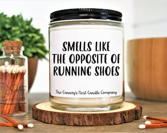 May include: A white candle jar with a black lid and a black label that reads "Smells like the opposite of running shoes" and "The Canary's Nest Candle Company".