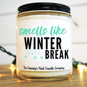 May include: A white candle jar with a black lid and a label that reads "smells like winter break". The label has a teal blue snowflake design and the text "The Canary's Nest Candle Company".