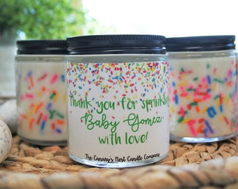 May include: Three glass candle jars with white wax and colorful sprinkles. The middle jar has a label that reads "Thank you for sprinkling Baby Gomez with love! The Canary's Nest Candle Company."