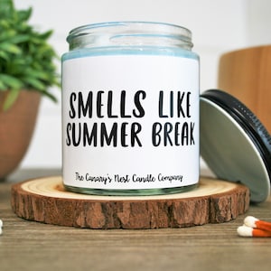 May include: A white candle jar with a black label that reads "Smells Like Summer Break" and "The Canary's Nest Candle Company".