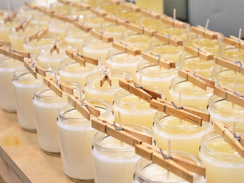 May include: A row of glass jars filled with white wax and a wick, each secured with a wooden clothespin. The jars are arranged in rows on a wooden surface.