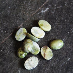 Puede incluir: Una colección de piedras ovaladas de color verde y blanco con un patrón moteado. Las piedras están dispuestas sobre una superficie gris oscura.