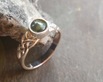 Può includere: Un anello in argento con una pietra cabochon rotonda verde scuro incastonata in una lunetta liscia. La fascia presenta un intricato design a nodo celtico traforato che si estende dall'incastonatura, creando un dettaglio metallico sui lati.