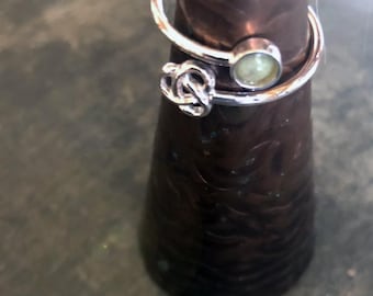 May include: A silver-colored ring with a knot design and a small, round, light green stone. The ring wraps around the finger with a curved design. The ring is displayed on a dark brown, cone-shaped object.