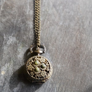 Puede incluir: Un colgante de reloj de bolsillo dorado con un delicado diseño floral y piedras preciosas verdes. El reloj está unido a una cadena dorada.