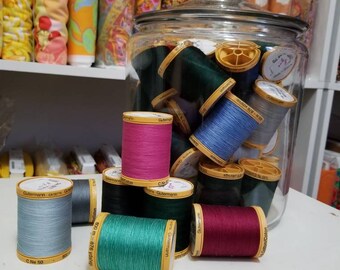 May include: A glass jar filled with spools of colorful thread, with several spools of thread in various colors, including blue, green, pink, red, and gray, sitting on a white surface. The spools of thread are labeled "Gutermann".