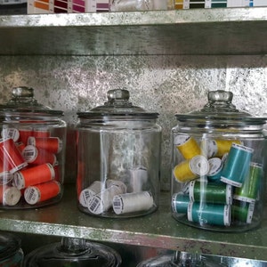 May include: Five glass jars filled with spools of colorful thread. The jars are arranged in a row on a shelf. The thread colors are purple, orange, white, green, and blue.