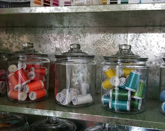 May include: Five glass jars filled with spools of colorful thread. The jars are arranged in a row on a shelf. The thread colors are purple, orange, white, green, and blue.