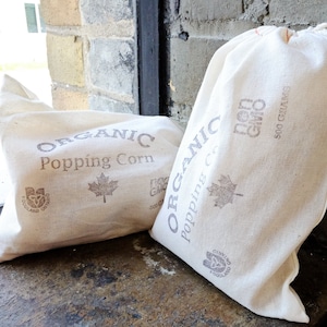May include: Two white canvas bags filled with organic popping corn. The bags have brown text that reads "Organic Popping Corn" and "Non GMO 500 Grams".