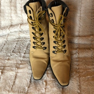 May include: A pair of tan ankle boots with black trim and yellow and black laces. The boots have a pointed toe and are set against a textured, light-colored background.
