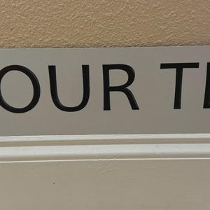 May include: A rectangular, silver-colored sign with the text "YOUR TEXT" in bold, black letters. The left side features a blue panel with a circle of yellow stars and the letter "D". The sign is angled slightly.