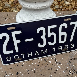 May include: A rectangular license plate with a navy blue background and white lettering. The plate reads "2F-3567 GOTHAM 1966". The plate has a white border and is displayed outdoors.