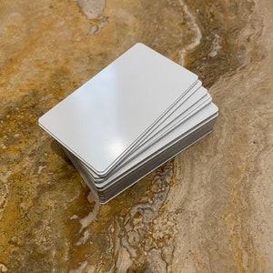 May include: A stack of blank white rectangular cards with rounded corners. The cards are stacked neatly, revealing their gray edges. The cards are on a brown and gold marbled surface.