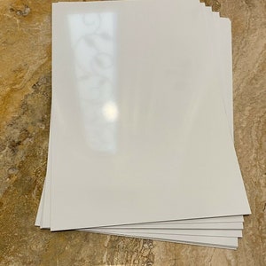 May include: A stack of white, glossy paper sheets. The paper has a smooth, reflective surface and is stacked neatly. The edges of the paper are clean and sharp. The paper is likely for printing or crafting.