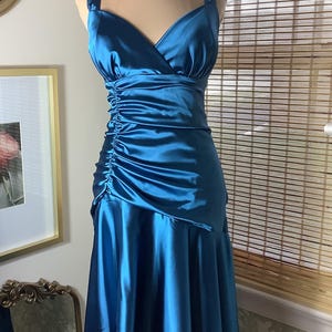 May include: A teal satin dress with a V-neckline and spaghetti straps. The bodice features ruching on the side, and the skirt has a flowing, asymmetrical hemline. The dress is displayed on a mannequin.