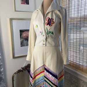 May include: A cream-colored two-piece outfit consisting of a long-sleeved jacket and a matching skirt. The jacket features a large embroidered flower with multi-colored petals. The skirt is decorated with a chevron pattern of colorful ribbons.