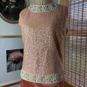 May include: A sleeveless, peach-colored top covered in sequins. The neckline and hem are trimmed with a beaded design in cream, gold, and peach. The top is displayed on a mannequin.