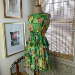 May include: A vintage, sleeveless dress with a floral pattern in shades of green, pink, and yellow. The dress features a tiered skirt and a yellow bow at the waist. It is displayed on a mannequin.