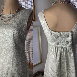 May include: A sleeveless, light silver dress with a subtle floral pattern. The dress features a square neckline and decorative buttons on the back. A multi-strand necklace is draped on the mannequin. The dress is displayed on a mannequin.
