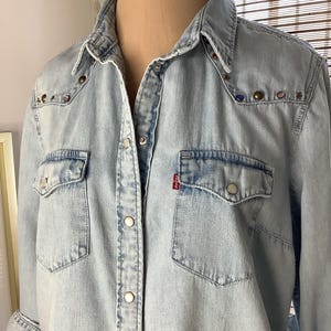 May include: Light blue denim shirt with two chest pockets and snap closures. The shirt features decorative studs on the shoulders and a small red tag on the pocket. A classic, casual top.