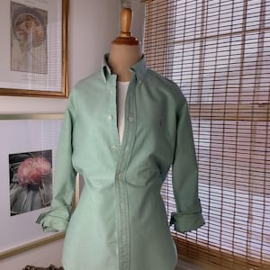 May include: A light green button-down shirt with long sleeves, displayed on a mannequin. The shirt has a classic collar and a small logo on the chest. The sleeves are rolled up. The background includes framed artwork and a bamboo blind.