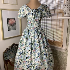 May include: A tea-length dress featuring a white base with a blue, green, and pink floral print. The garment has short, ruffled sleeves, a V-neckline, and a fabric flower accent at the center of the bodice. The skirt is full and gathered at the waist.