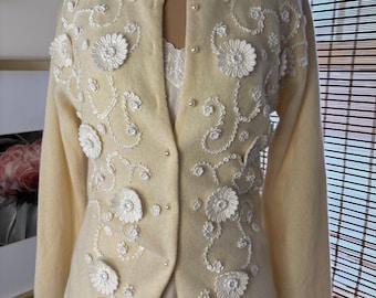 Puede incluir: Un cárdigan amarillo pálido adornado con bordados florales blancos y botones perlados. El suéter tiene un cuello redondo y mangas largas. El diseño floral se concentra en la parte delantera. Estilo vintage.
