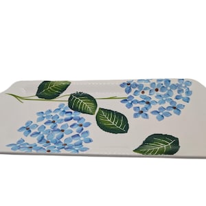 May include: A rectangular white ceramic serving platter with a hand-painted design of blue hydrangea flowers and green leaves. The platter has a slightly curved shape and is suitable for serving food.