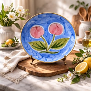 May include: A hand-painted ceramic plate with a blue background and a gold rim. The plate features two pink flower designs with green leaves. The plate is displayed on a wooden cutting board with a brass stand.