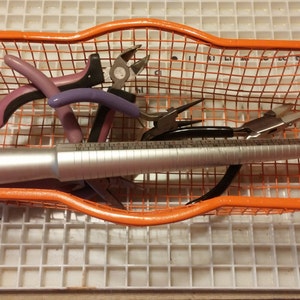 May include: An orange wire basket filled with jewelry-making tools. The tools include pliers with purple and black handles, a ring sizer, and other metal tools. The basket sits on a white grid surface.