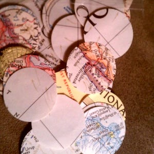 May include: A collection of circular paper cutouts, each featuring sections of vintage maps. The maps display various colors and geographical details. Some circles are blank with a diagonal line. The overall theme is travel and geography.