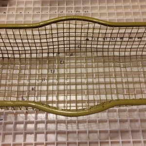 May include: An olive green wire mesh basket with a rounded rectangular shape. The basket is made of a grid of thin wires and has a solid, curved border. The basket is empty and sits on a white grid surface.
