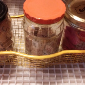 May include: Three glass jars with lids sit in a yellow wire basket. The jars contain small wooden letter tiles and pink items. The jars have black, orange, and gold lids. The basket sits on a white grid surface.