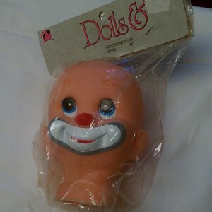 May include: A vintage doll head in a plastic bag. The head is peach-colored with blue eyes, a red nose, and a white and gray clown smile. The packaging reads "Dolls &" and "HOBO HEAD 4 1/2 IN".
