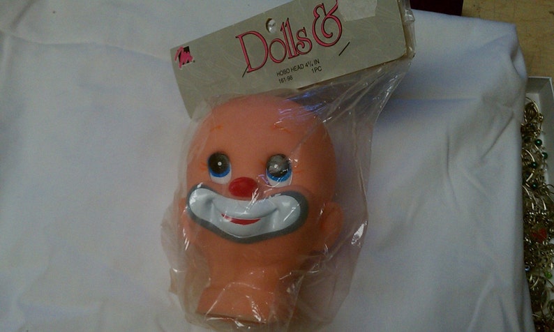 May include: A vintage doll head in a plastic bag. The head is peach-colored with blue eyes, a red nose, and a white and gray clown smile. The packaging reads "Dolls &" and "HOBO HEAD 4 1/2 IN".