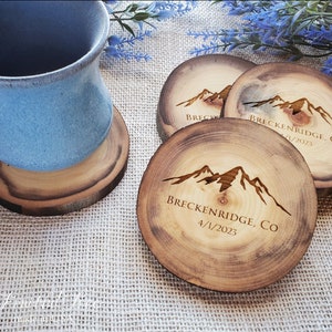 May include: A set of round wooden coasters with natural bark edges, each featuring a laser-engraved mountain range graphic and the text "BRECKENRIDGE, CO" followed by the date "4/1/2023". One coaster supports a light blue ceramic mug.