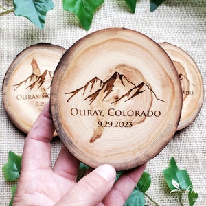 May include: Three circular wooden coasters with natural bark edges. Each coaster features a dark brown laser-engraved mountain range graphic above the text "OURAY, COLORADO" and the date "9.29.2023" centered on the light tan wood grain surface.