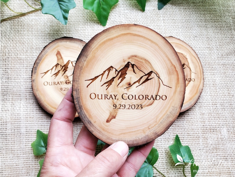 May include: Three circular wooden coasters with natural bark edges. Each coaster features a dark brown laser-engraved mountain range graphic above the text "OURAY, COLORADO" and the date "9.29.2023" centered on the light tan wood grain surface.