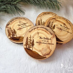 May include: Four circular wooden coasters with natural bark edges, each laser-engraved with a pine tree forest graphic, the text "Thank you for celebrating with us," "The Bolducs," "09-26-26," and "Lake Arrowhead, California" in dark brown ink.