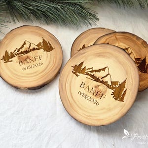 May include: Three round wooden coasters with natural bark edges, each featuring a laser-engraved mountain range and pine tree landscape. The text BANFF and the date 6/18/2026 are engraved below the mountain graphic on each light brown wood slice.
