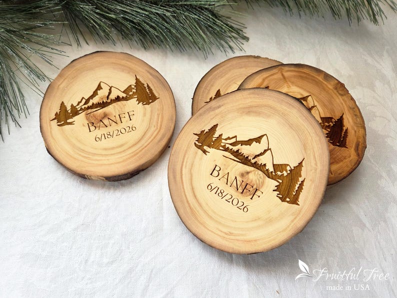 May include: Three round wooden coasters with natural bark edges, each featuring a laser-engraved mountain range and pine tree landscape. The text BANFF and the date 6/18/2026 are engraved below the mountain graphic on each light brown wood slice.