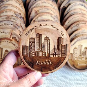 May include: A set of round wooden coasters with a laser-engraved design of the Boston skyline. The coasters are personalized with the names "Maddie" and "Jaime" and the text "Hampshire House Boston, MA".