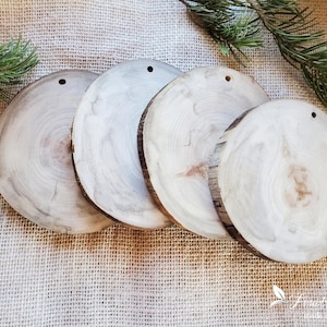 May include: Four circular wooden slices with natural bark edges and visible grain patterns. Each light-colored wood disc features a small pre-drilled hole near the top edge for hanging. The surfaces are smooth, showing concentric growth rings.