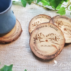 May include: Three round wood slice coasters featuring a laser-engraved mountain landscape, the names Jenna & Austin, the date 09.29.2023, and the text Thank you for celebrating with us! One coaster supports a blue ceramic mug.