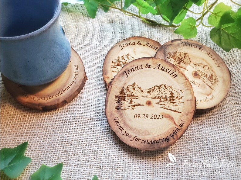 May include: Three round wood slice coasters featuring a laser-engraved mountain landscape, the names Jenna & Austin, the date 09.29.2023, and the text Thank you for celebrating with us! One coaster supports a blue ceramic mug.