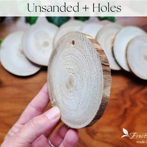 May include: A close-up of a pile of unfinished wood slices with holes drilled in the centre. The wood is a light brown colour and has a natural, rustic look. The slices are arranged in a pile, with the top slice being held in a hand.