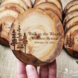 May include: A round wooden coaster featuring a natural bark edge and a laser-engraved design of pine trees. The text reads Walk in the Woods Wellness Retreat and February 10, 2024. The wood grain is visible, showing light brown rings and dark edges.