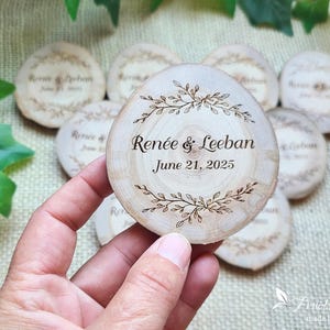 May include: A round wooden slice featuring a laser-engraved design of leafy branches framing the text Renée & Leeban and the date June 21, 2025. The surface shows natural wood grain patterns and light bark edges, with multiple identical pieces visible.
